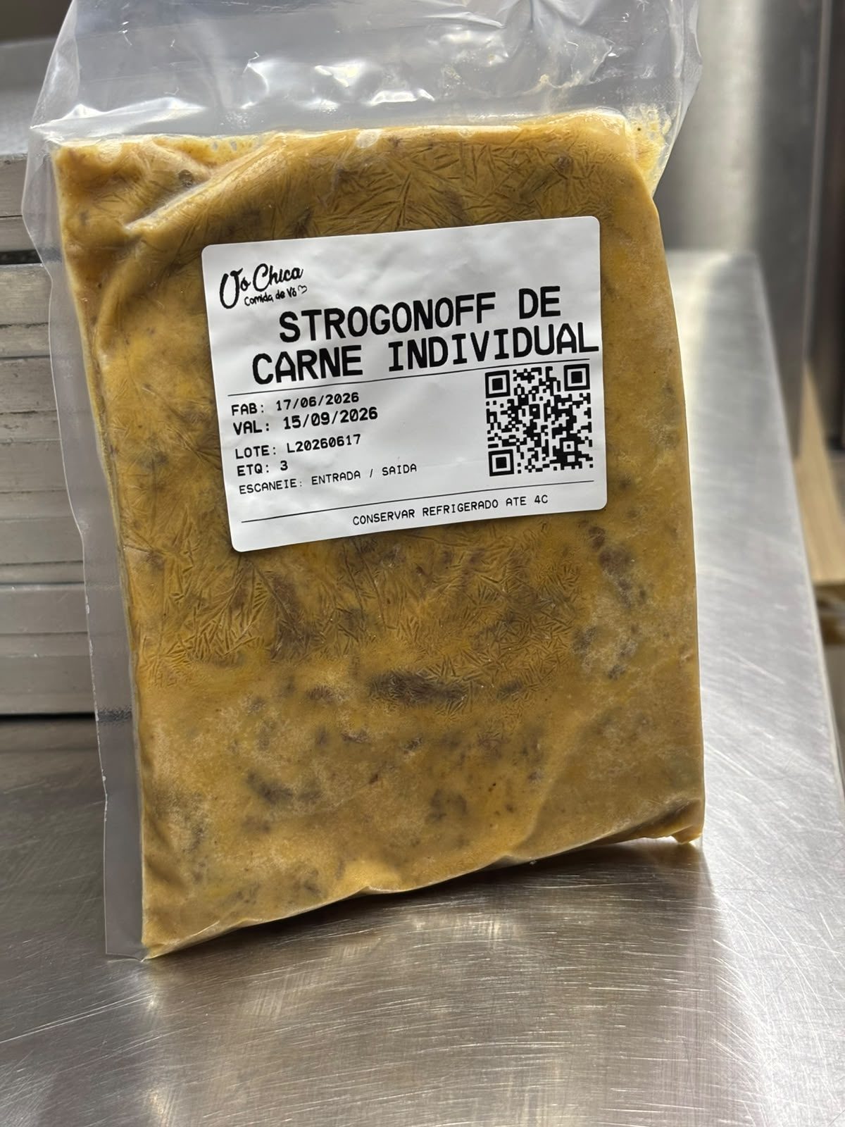 Etiqueta impressa da Vó Chica colada num saco a vácuo de comida congelada sobre a bancada de inox: nome do produto Strogonoff de Carne Individual em letra grande, data de fabricação, validade, lote, número da etiqueta, QR code para entrada e saída e a instrução de conservar refrigerado até 4 graus.