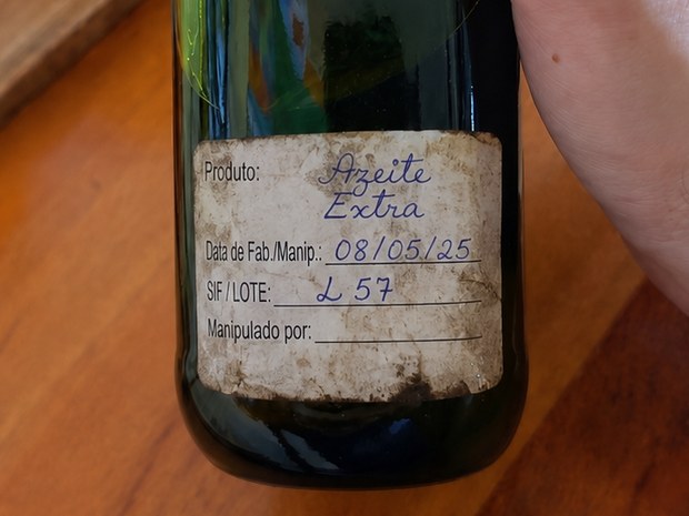 Etiqueta manuscrita e encardida colada numa garrafa de azeite: produto e data de fabricação preenchidos à caneta, campo SIF/Lote com uma anotação curta e o campo Manipulado por completamente em branco. Não há campo de validade.